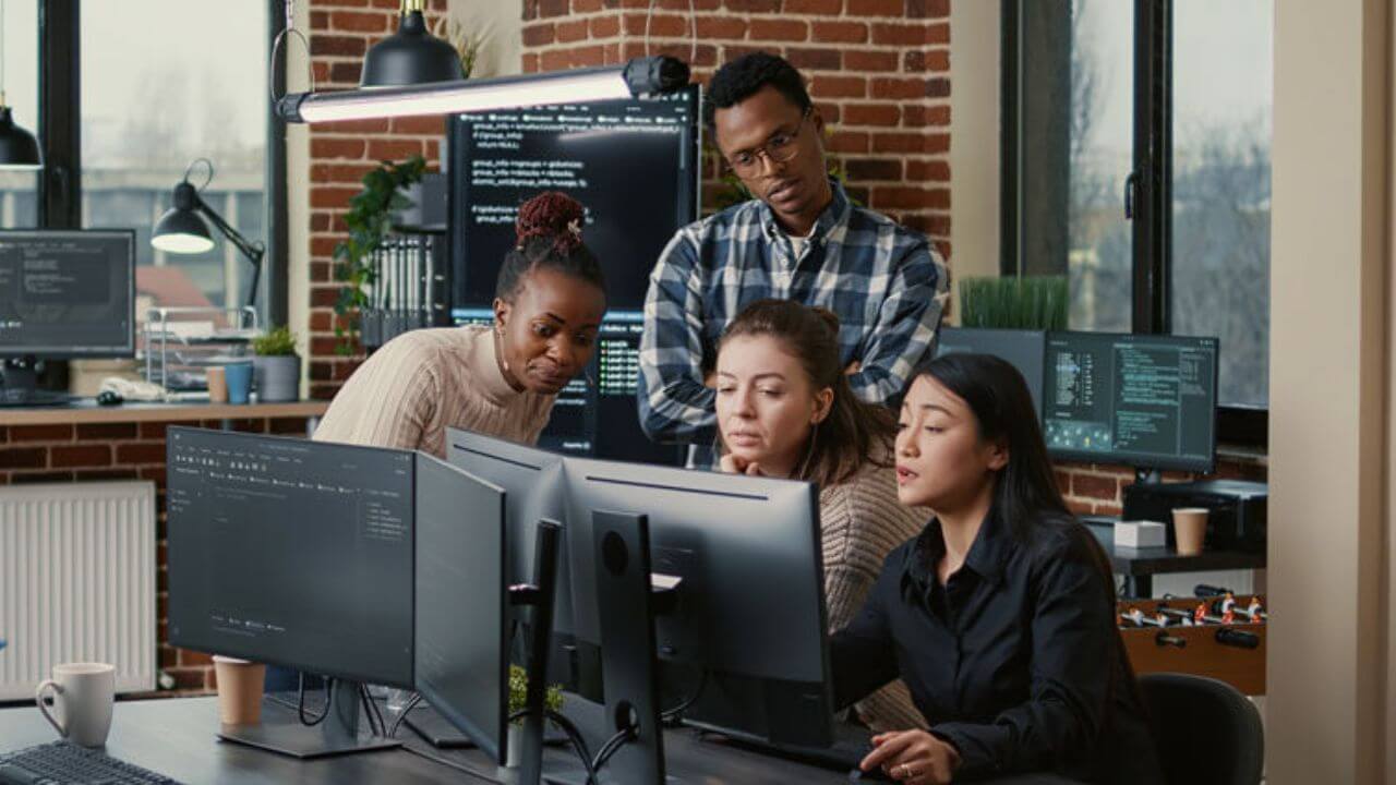 Software engineers brainstorming ideas looking at code on computer screens asking feedback from senior developer while intern joins the discussion. Junior programmers collaborating on group project.